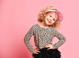 Portrait of playful blonde curly kid girl in leopard print sweatshirt, black skirt and fashion visor cap dancing