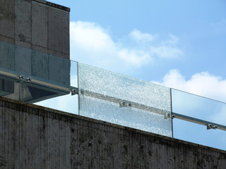 Shattered glass balustrade railing. broken laminated tempered safety glass. stone panel facade. construction and building industry concept. stainless steel glass bracket spiders. blue sky white clouds - obrazy, fototapety, plakaty