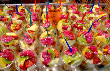 fruit cups of freshly sliced fruit mixture in plastic cups with lid and colorful forks in a fruit market in day light. isolated. displayed tastefully in bright light. healthy eating concept