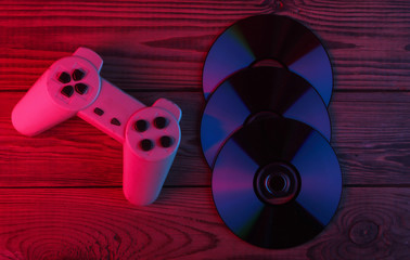 CD discs, gamepad on wooden background. Neon red blue light. Gradient glow. Retro media and...