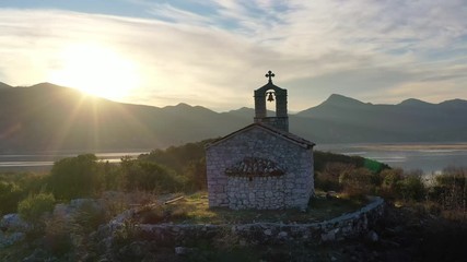 Small orthodox church made of stone, on the top of a hill, by Lake Skadar in Montenegro - Silhouette against beautiful sunset sky. Aerial footage.