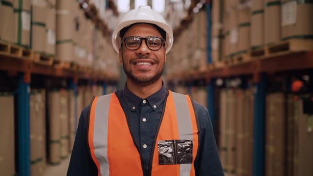 Tilt up short of a smiling young male warehouse worker or a supervisor standing in a warehouse with his arms crossed looking at camera