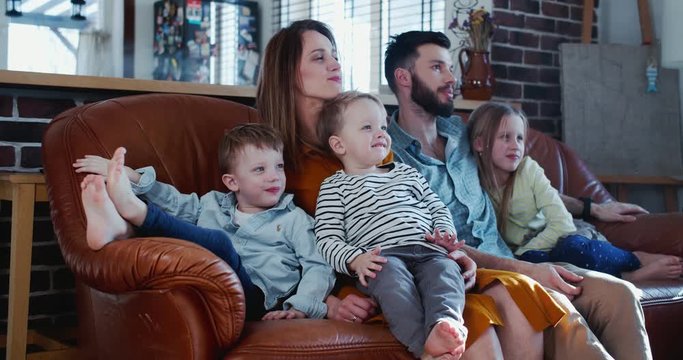 Family time together concept, happy young Caucasian parents and three children watch TV at home on the couch slow motion