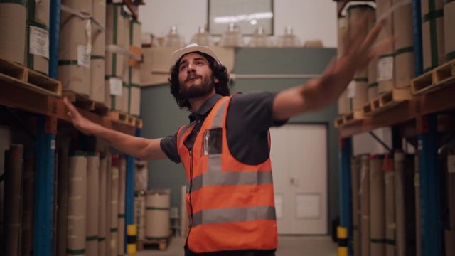 Carefree joyful young male worker in white hardhat and reflective jacket dancing between the rack inside the warehouse