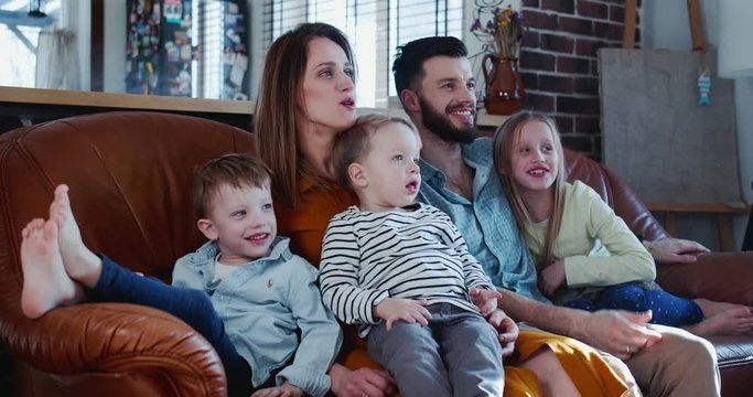 Cheerful happy young Caucasian family with three cute children laugh watching movies at home on the couch slow motion.