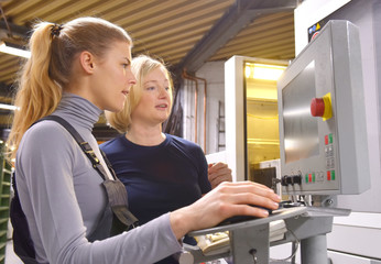 A young trainee receives tuition from her trainer. Both women work as computerized  numerical control engineers. They are seen at their high tech place of work.