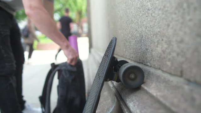 White male in black pants leans a black skateboard against a marble wall in a park and then takes off his backpack.