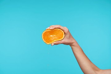 Beautiful fair-skinned pretty hand keeping her hand raised while holding fresh orange, squeezing it with fingers while isolated against blue background