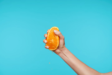 Colorful shot of raised female's hand with half of fresh orange, smashing it with fingers while...