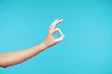 Side view of young woman's raised hand with white paint on it forming fingers in ok gesture, showing well done sign while isolated over blue background