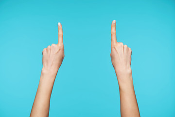 Photo of two raised hands of young woman with white manicure keeping up index fingers while pointing upwards, standing against blue background