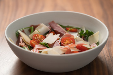 Salad with prosciutto, tomato, arugula leaves and parmesan cheese in white bowl on walnut table