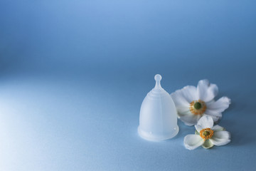 Obraz na płótnie Canvas Menstrual cup with white flowers on pastel blue background. Gentle care female health and eco concept, hygiene period product. soft light