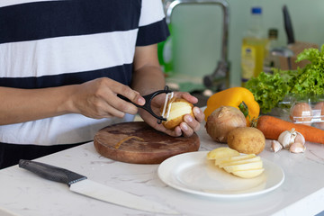 man peeling and cutting a potato on a white kitchen table with vegetables in the background