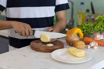 man cutting a potato on a white kitchen table with vegetables in the background
