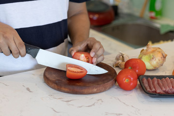 man cutting a tomato white a knife on a white kitchen table with vegetables in the background