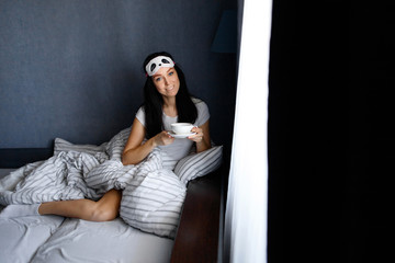 Happy beautiful woman in a sleep mask sitting on the bed in the house with a Cup of coffee/tea