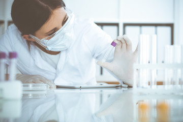 Professional female scientist in protective eyeglasses and mask making experiment with reagents in...