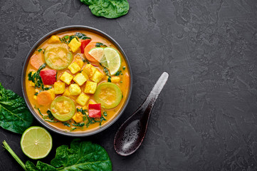 Yellow Veg Thai Curry with Tofu and vegetables in black bowl at dark slate background. Vegetarian Thai Curry with tofu, zucchini, pepper, spinach, carrot. Thai Food. Indian vegetable curry. Copy space - obrazy, fototapety, plakaty