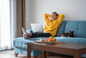 Happy young woman in a yellow sweatshirt works at home on a blue sofa with a laptop and a cat,...