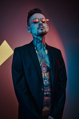 Cool male model posing in a neon studio wearing a black tuxedo on a half-naked body, fire-shaped sunglasses and tattooed in a japanese irezumi style, looking bold and calm. Vertical portrait