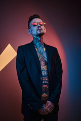 Bold and rebellious male model posing in a neon studio wearing a black tuxedo on a half-naked body, fire-shaped sunglasses and tattooed in a japanese irezumi style, looking self-assured. Vertical