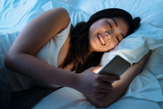 technology internet communication and people concept. happy smiling young woman text online message on smartphone in white bed at home bedroom at night. laughing girl using cellphone lying on pillow
