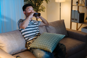 Fototapeta premium Young asian chinese man watching television drinking bottle of beer in late evening time. handsome guy enjoy alcohol alone in sofa at night in apartment. single male stay up late relax on couch