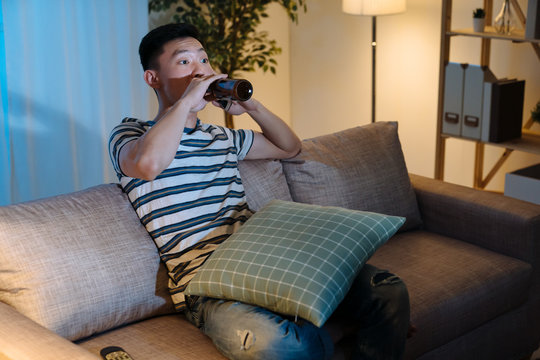 Young Asian Chinese Man Watching Television Drinking Bottle Of Beer In Late Evening Time. Handsome Guy Enjoy Alcohol Alone In Sofa At Night In Apartment. Single Male Stay Up Late Relax On Couch