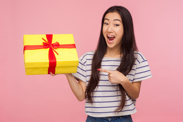Awesome birthday present! Amazed satisfied pretty girl with brunette hair in striped t-shirt pointing at gift box and looking with surprised expression. indoor studio shot isolated on pink background