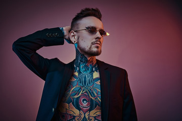Bold and rebellious male model posing in a neon studio wearing a black tuxedo on a half-naked body,...
