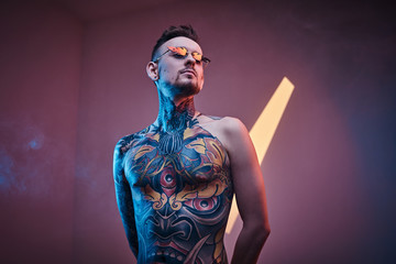 Self-assured male model posing in a neon studio with a half-naked body wearing fire-shaped sunglasses and tattooed in a japanese irezumi style, looking cool and relaxed.