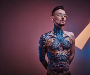 Assertive male model posing in a neon studio with a half-naked body wearing sunglasses and tattooed...