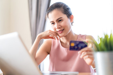 Asian young woman sit in chair, while using laptop for shopping online  from home. Girl hold credit card, smile and wait for delivery package box by shipping company. Shopaholic Lifestyle concept.