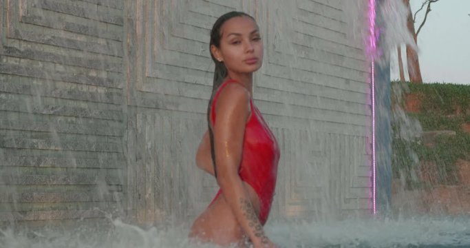 Beautiful brunette woman in red swimsuit posing in outdoor pool over wall with waterfall background - video in slow motion