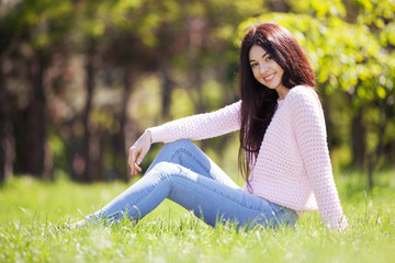 Young woman relax in the park on green grass. Beauty nature scene with colorful background, trees at spring season. Outdoor lifestyle. Happy smiling woman sitting on green grass