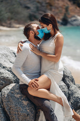 Fototapeta na wymiar Portrait of a beautiful couple kissing sexually on the beach next to the azure sea while wearing a face mask that protects them from infection because of the outbreak of coronovirus infection COVID-19