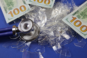 Health care system collapse concept: us dollar money bill notes on pile of broken glass shards, stethoscope, blue background (focus on stethoscope)