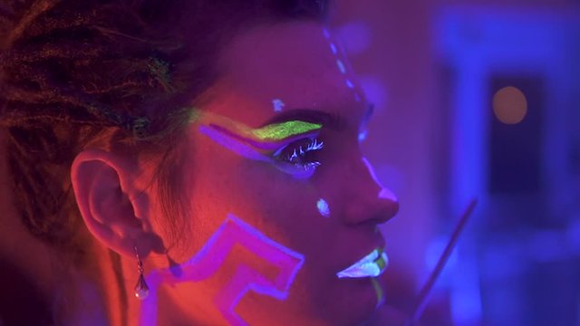 Making up a Girl with Dreadlocks in Neon UF Light. Model Girl with Fluorescent Creative Psychedelic MakeUp, Art Design of Female Disco Dancer Model in UV, Colorful Abstract Make-Up. Dancing Lady