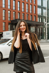 Happy pretty girl in formal casual clothes talking on the phone in the city street with a smile on her face and looking into the camera. Cheerful businesswoman talking on the phone, street portrait