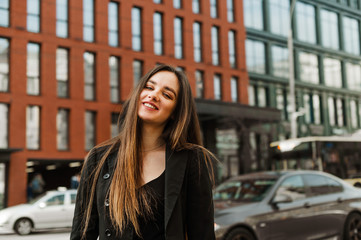 Cheerful young businesswoman in dark formal wear posing at camera on cityscape background, standing on street against modern architecture background. Happy lady in jacket smiling at camera.