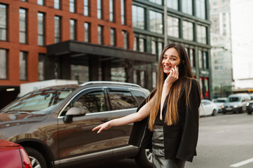Fototapeta na wymiar Positive girl in formal clothes stands on the road and catches a taxi with her hand raised. Cheerful young businesswoman in dark clothing is calling on the phone and hand gesture stops a taxi.