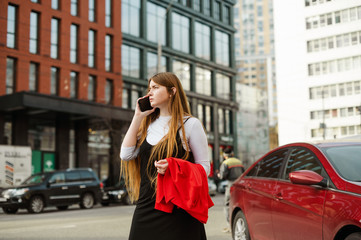 Portrait of a serious long-haired student girl standing on the street of a metropolis and talking on the phone against a background of cityscape. Student girl stands on the road and calls in a taxi.