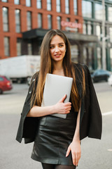 Pretty lady in casual casual clothes walking with a smile on her face down the street of a metropolis with a laptop in her hands on wireless headphones, looking away. Vertical portrait businesswoman.
