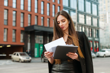 Business girl in wireless headphones stands with laptop and business papers against a backdrop of a street landscape and reads information on documents with a serious face. Business people concept.