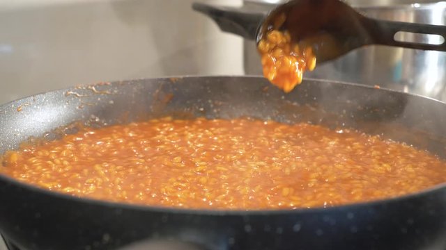 Cooking risotto rice with tomato sauce - close up of female hands and skillet on the burner cooking and pouring broth on the rice and mixing - Italian typical recipe