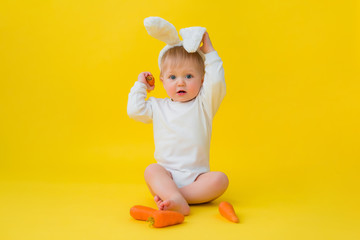 Baby in a white bodysuit with rabbit ears on his head eats carrots, sits on a yellow background with vegetables. baby in the form of an Easter bunny, space for text