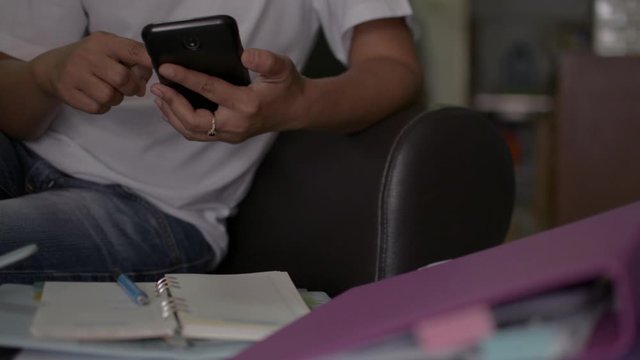 Woman in casual dress sitting on a leather sofa using mobile phone and working with a pile of document at home during COVID-19 outbreak situation. Work from home concept. Panning shot.