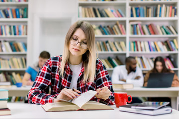 Young girl student, wearing casual checkered shirt and eyeglasses, sitting at the table in library and reading a book, preparing for test or exam. Her groupmates studying on the background