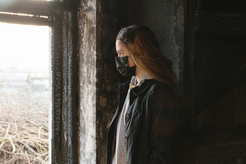 A teenager girl in a black medical mask looks out the window from an abandoned post-apocalyptic building. Consequences of COVID-19 Coronavirus protection. After a pandemic
